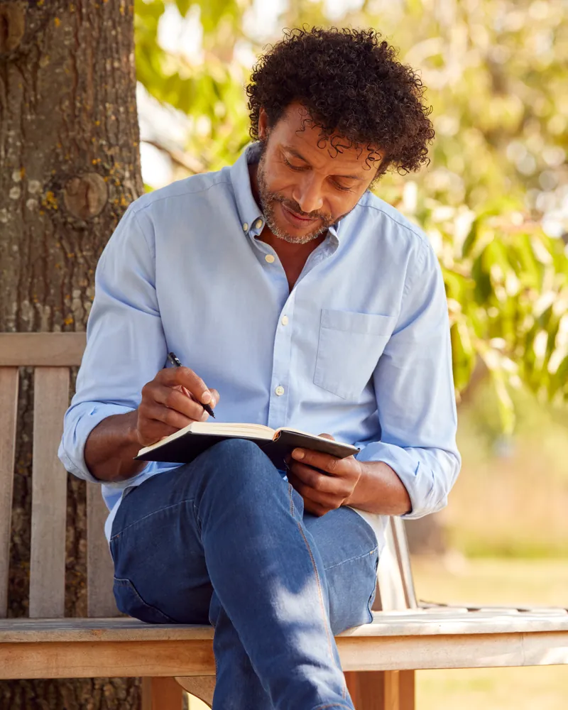 A man writes in a notebook on a park bench under a tree